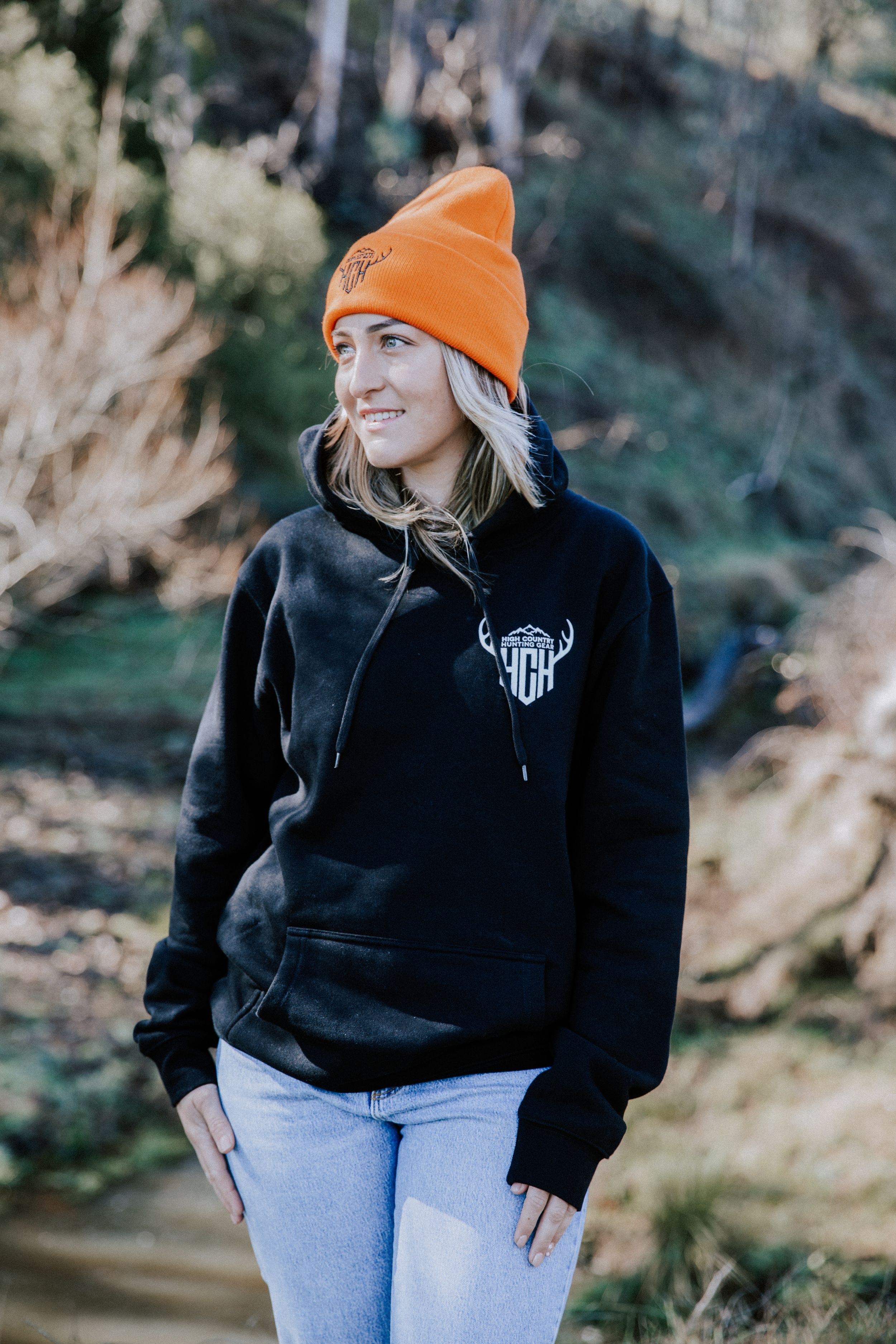 Someone wearing an orange beanie and black hoodie with an antler logo stands outdoors near water in autumn.