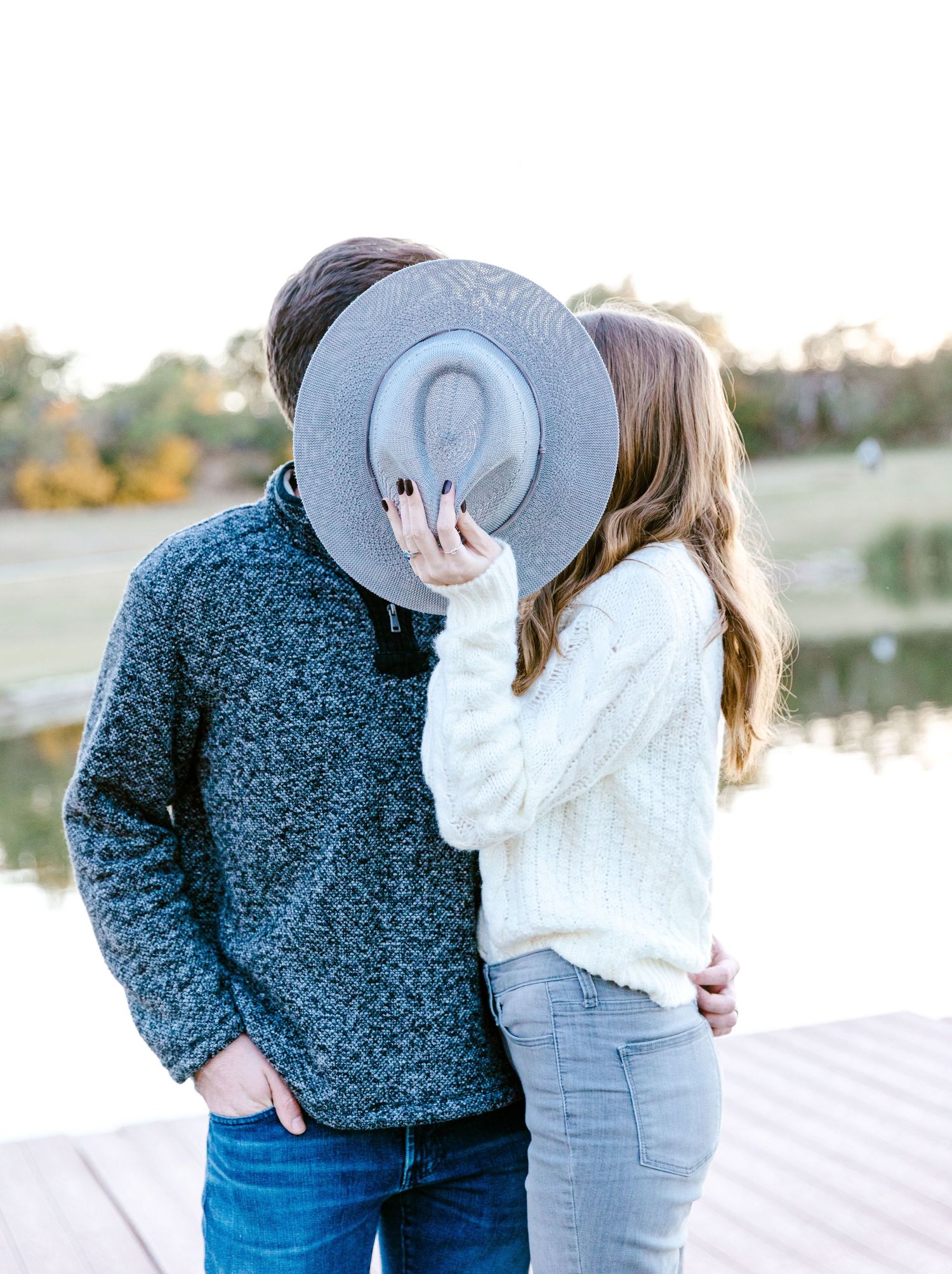 A romantic couple sharing an intimate moment by a lake with a gray hat covering their faces.