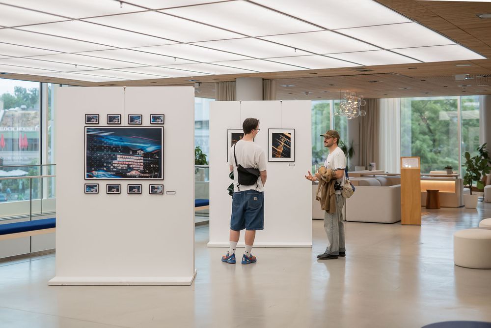 Two visitors view framed photographs on white display walls in a bright, modern gallery with large windows and lounge seating.