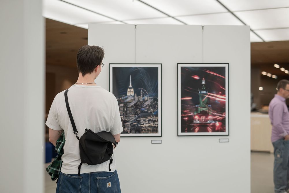 Visitor with a crossbody bag viewing two framed night cityscape photographs on a white wall in a bright modern art gallery.