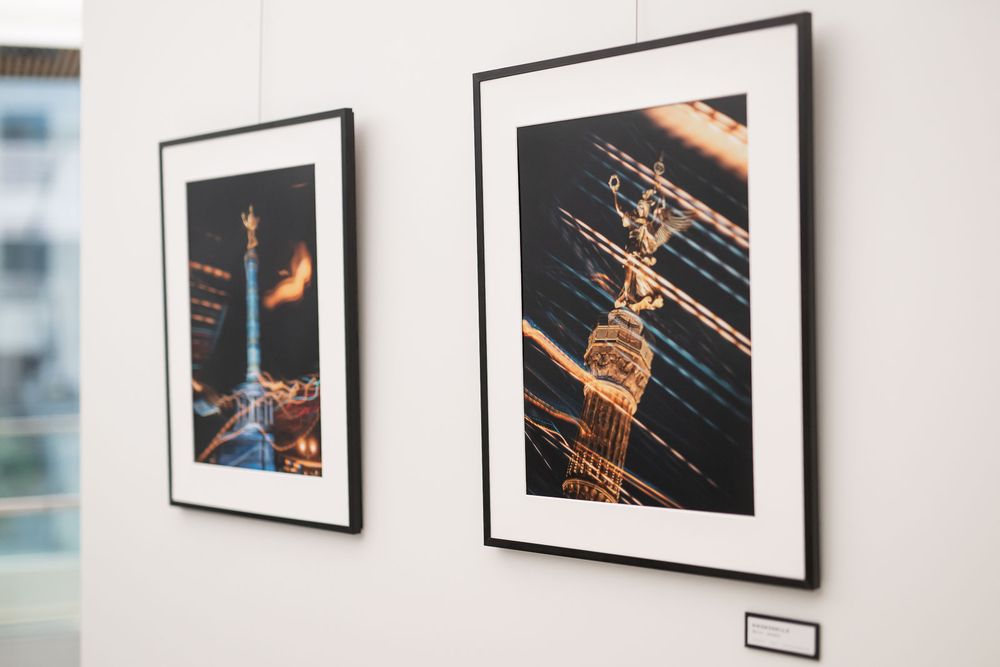 Two framed night photographs of an illuminated golden monument column hang on a white gallery wall beside a small label.