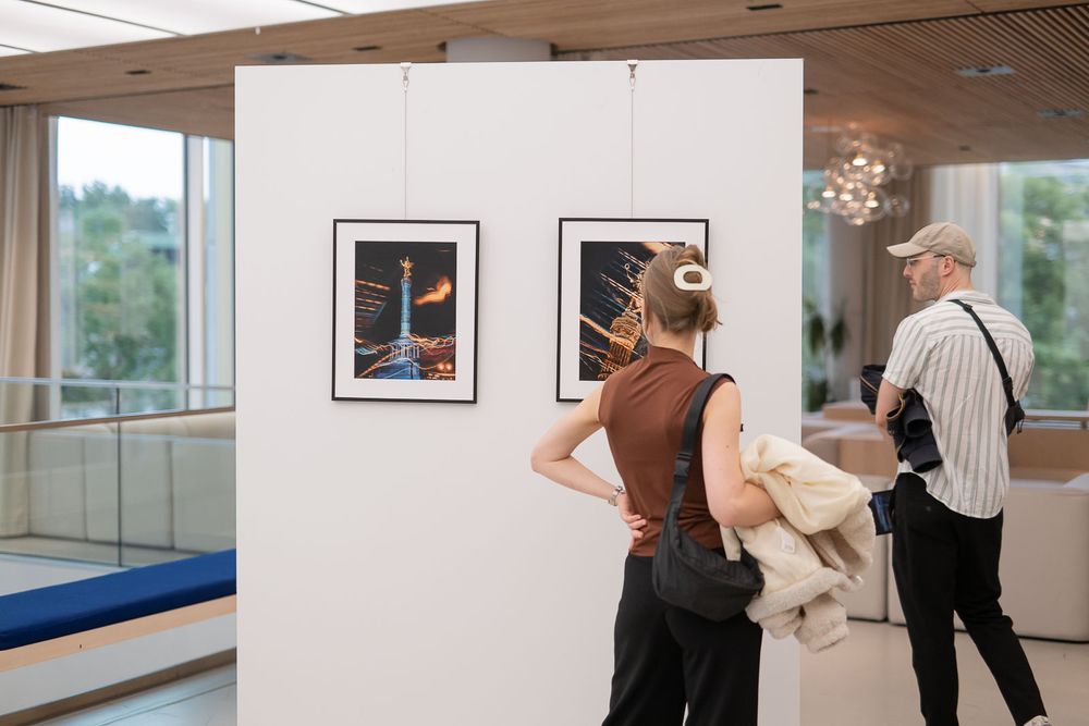 Visitors view framed night photographs on a white gallery partition in a bright modern exhibition space with large windows.