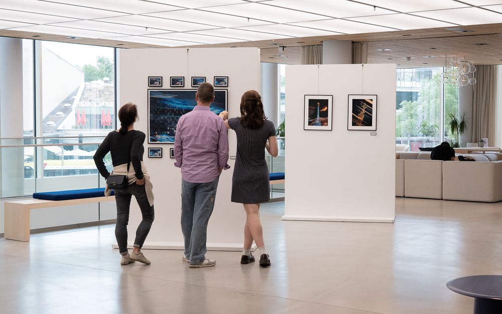 Three visitors view framed photographs displayed on white partition walls in a bright modern gallery with large windows and a blue bench.