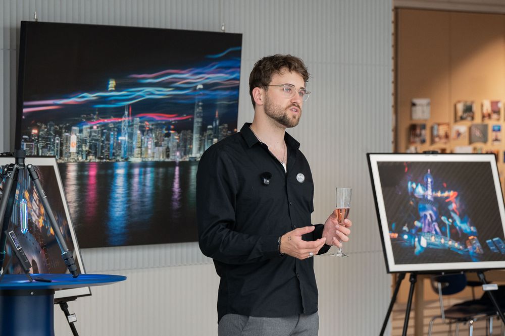 Bearded man in glasses and a dark shirt speaks while holding a glass, beside framed night cityscape photographs on display.