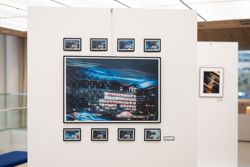 Large framed nighttime photograph of an illuminated building surrounded by smaller prints on a white gallery wall in a modern exhibition.
