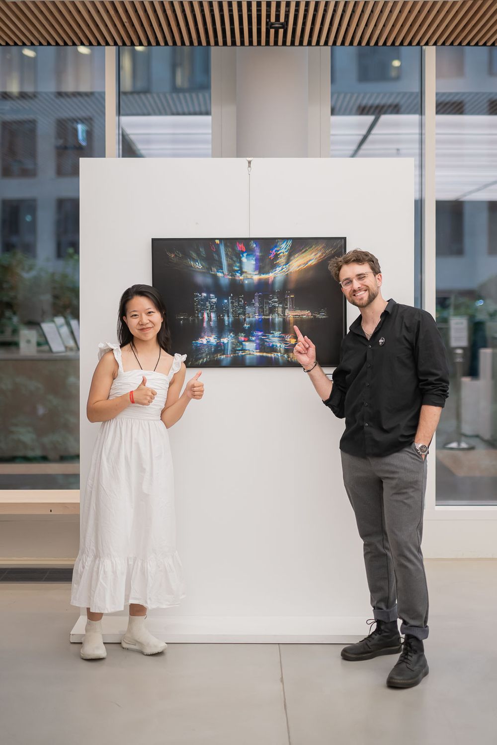 Two smiling people pose beside a framed night cityscape photograph on a white gallery wall, one giving thumbs up and the other pointing.
