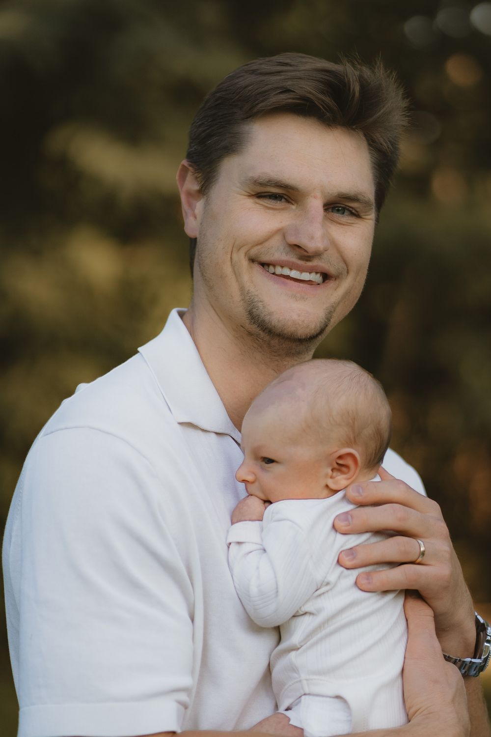 Smiling man in a white polo shirt gently holds a sleeping newborn baby in a white outfit outdoors with a soft, blurred green background.
