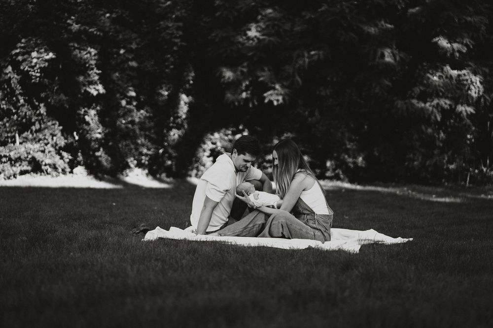 Black and white photo of a couple sitting on a blanket on a grassy lawn with tall trees in the background.