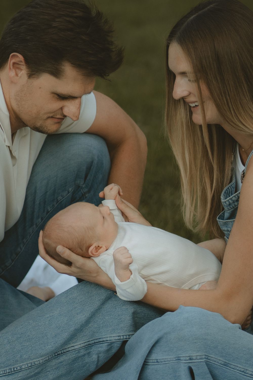Smiling parents in denim outfits gently cradle a sleeping newborn baby in a white bodysuit while sitting on the grass.