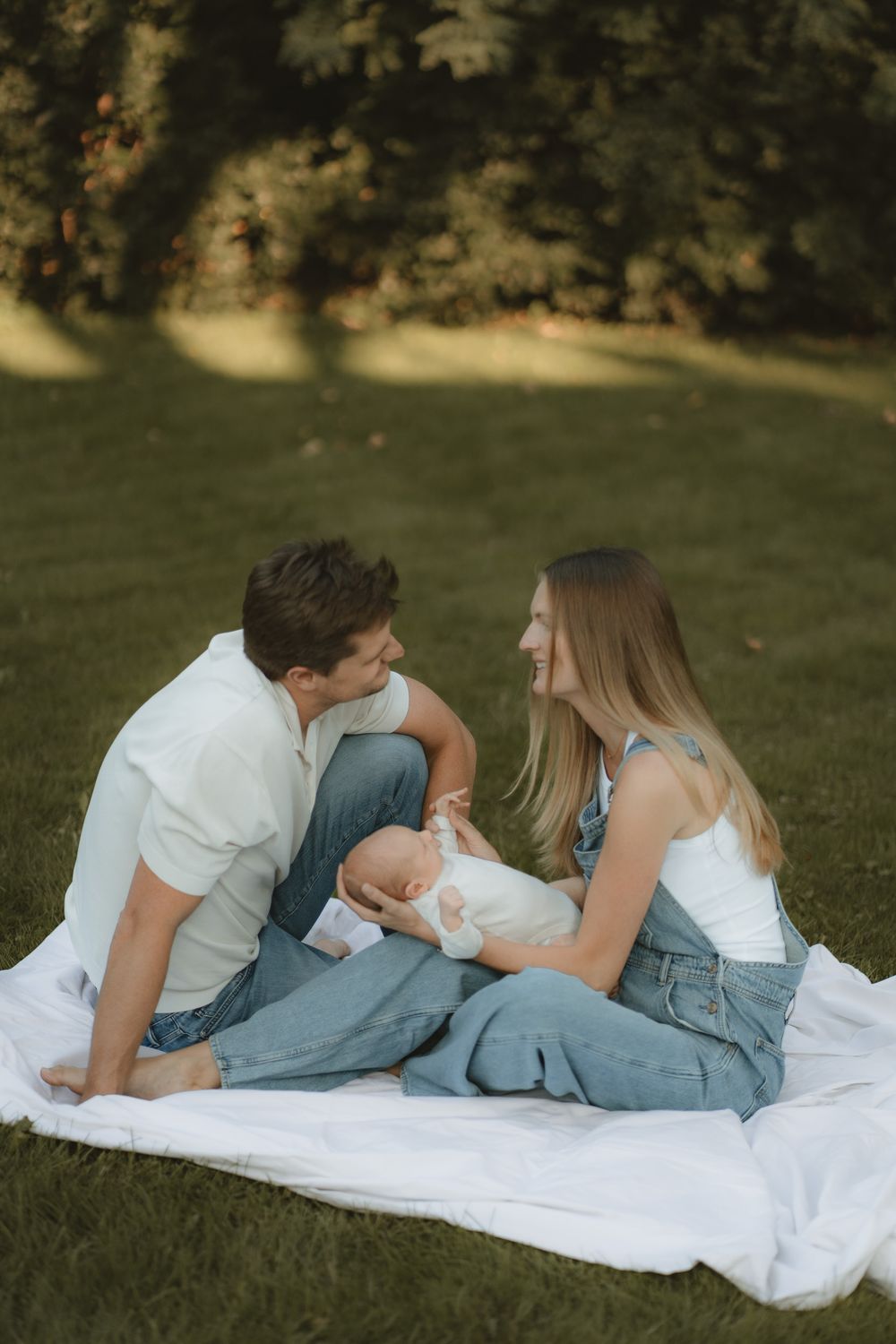 Smiling couple in casual clothes sit on a white blanket in a sunlit park, gazing lovingly at each other while holding their newborn baby.