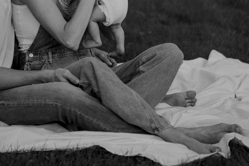Black and white photo of a barefoot couple in jeans sitting close together on a white blanket in the grass, embracing a small child.