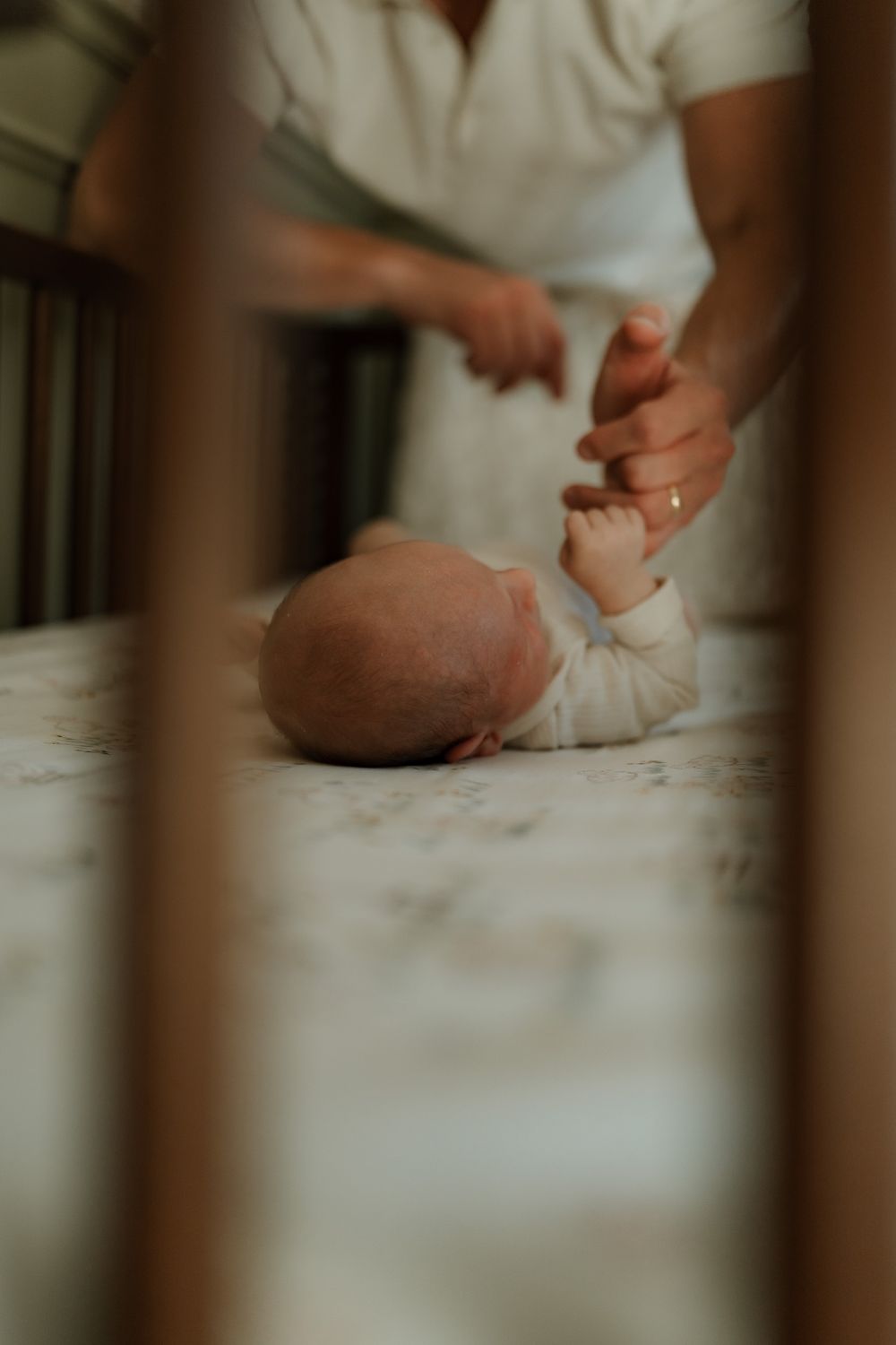 Baby lying on a patterned crib mattress holding an adult's hand, seen through wooden crib slats in soft natural light.