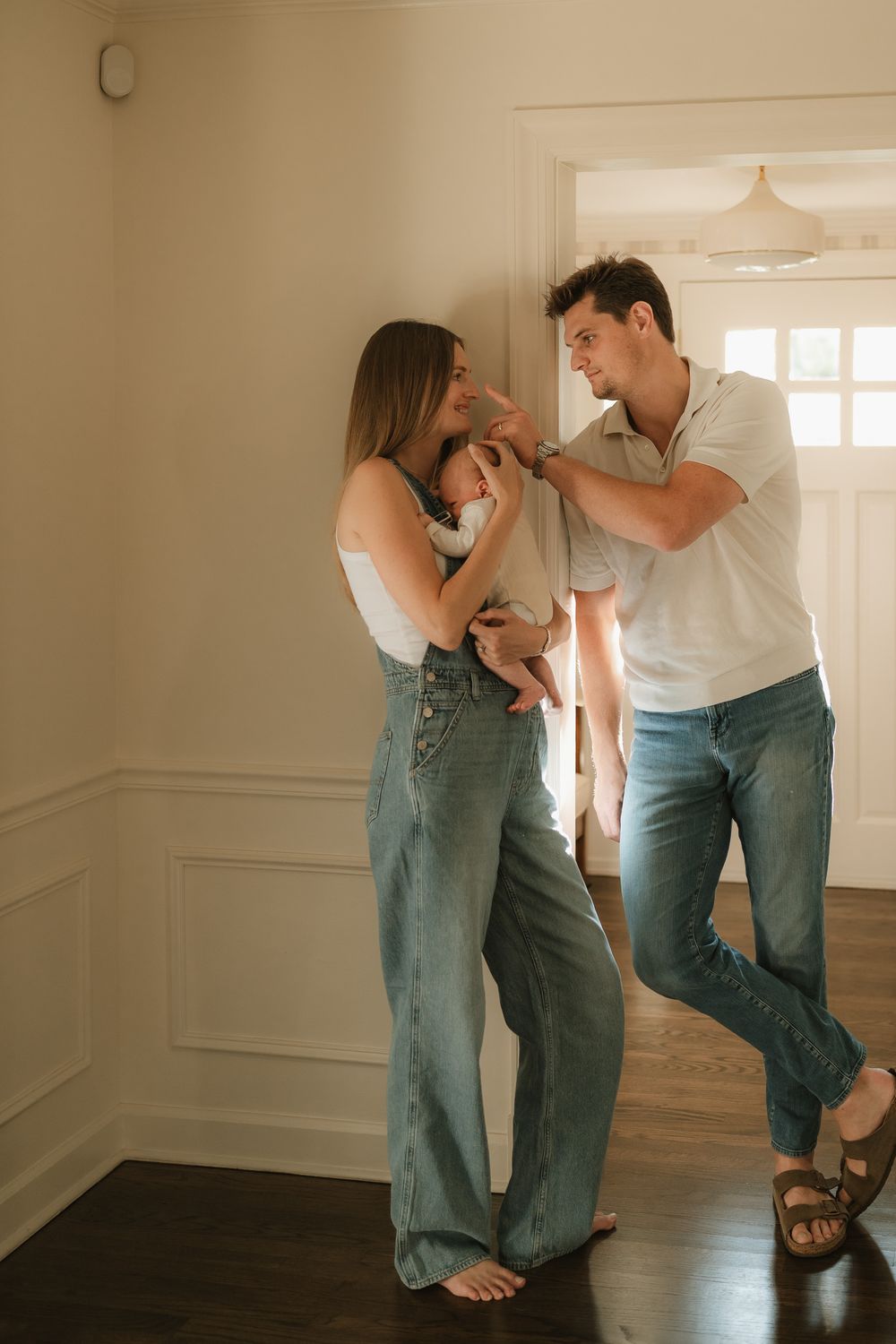 A smiling couple in denim stands in a bright hallway, gently cradling a newborn baby while the father touches the infant's head.
