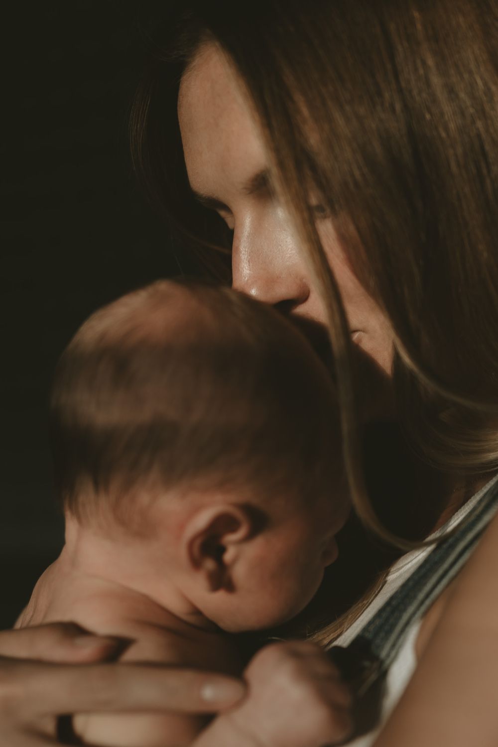 Mother with long brown hair gently kisses her sleeping newborn baby's head in soft, warm, moody light.