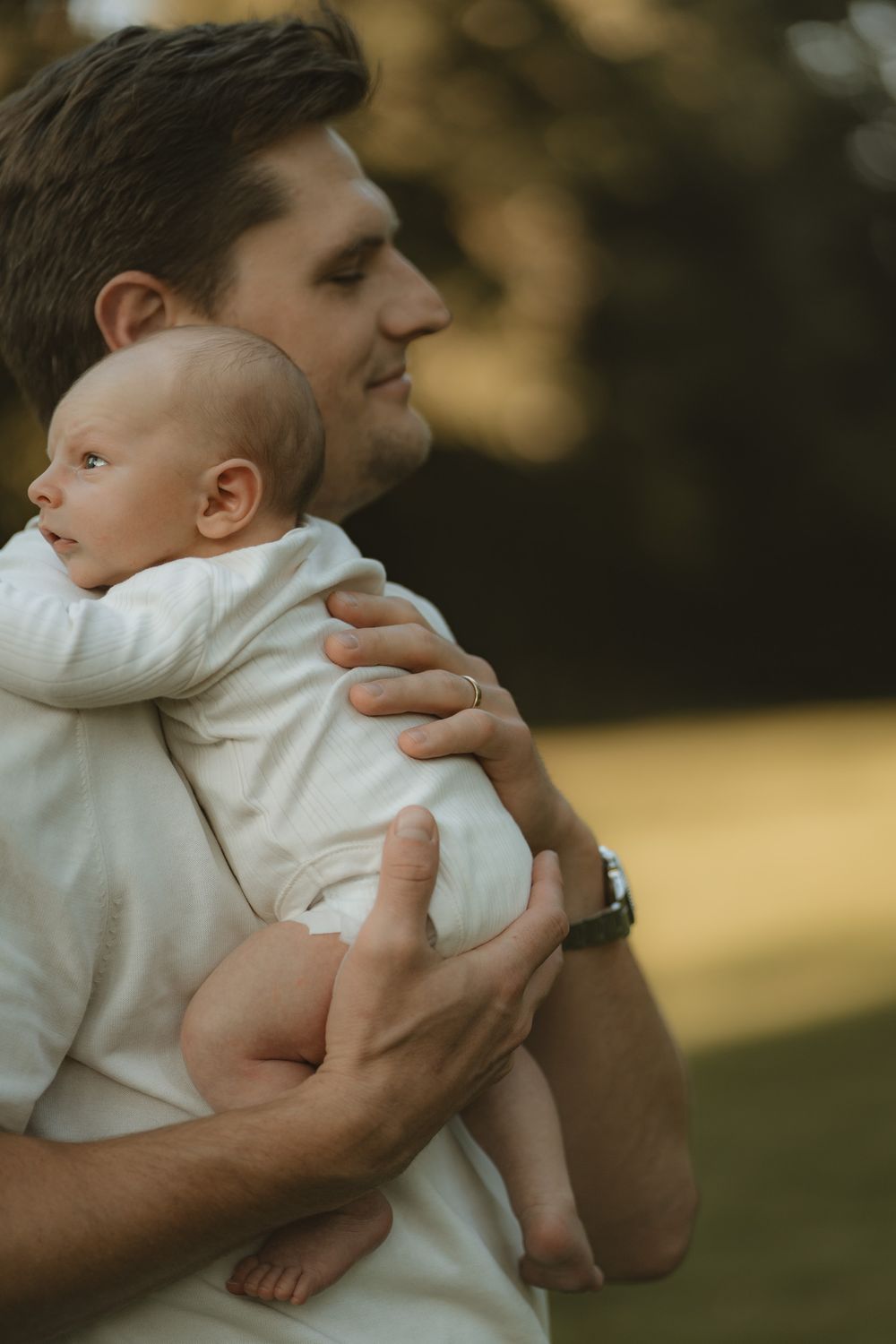 A smiling father in a white shirt holds a sleepy newborn baby in a cream outfit against his shoulder in a sunlit green park in Lake Forest