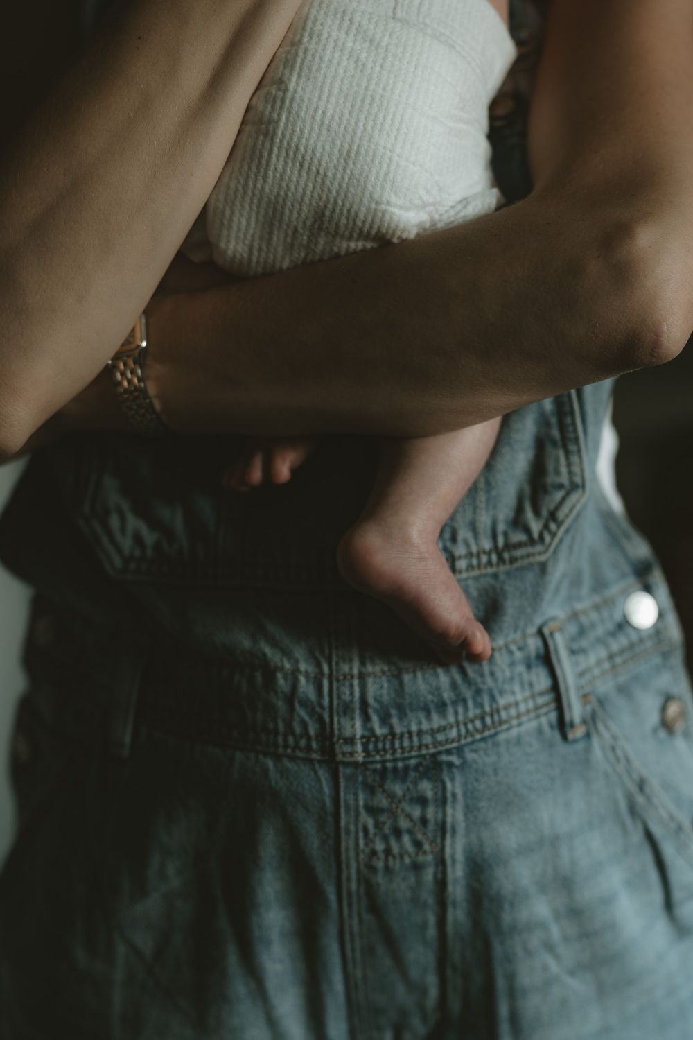 Close-up of a mother in denim overalls cradling a baby in a diaper, with the infant's bare foot visible.