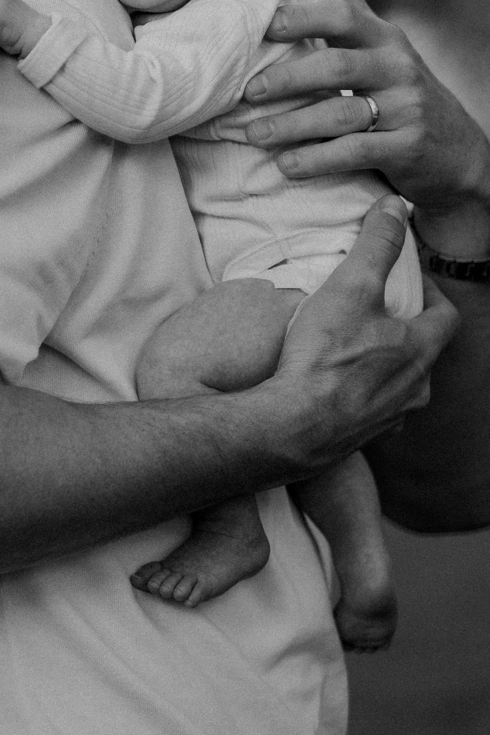 Black and white close-up of a father's hands wearing a ring, gently cradling a newborn baby with tiny bare feet.