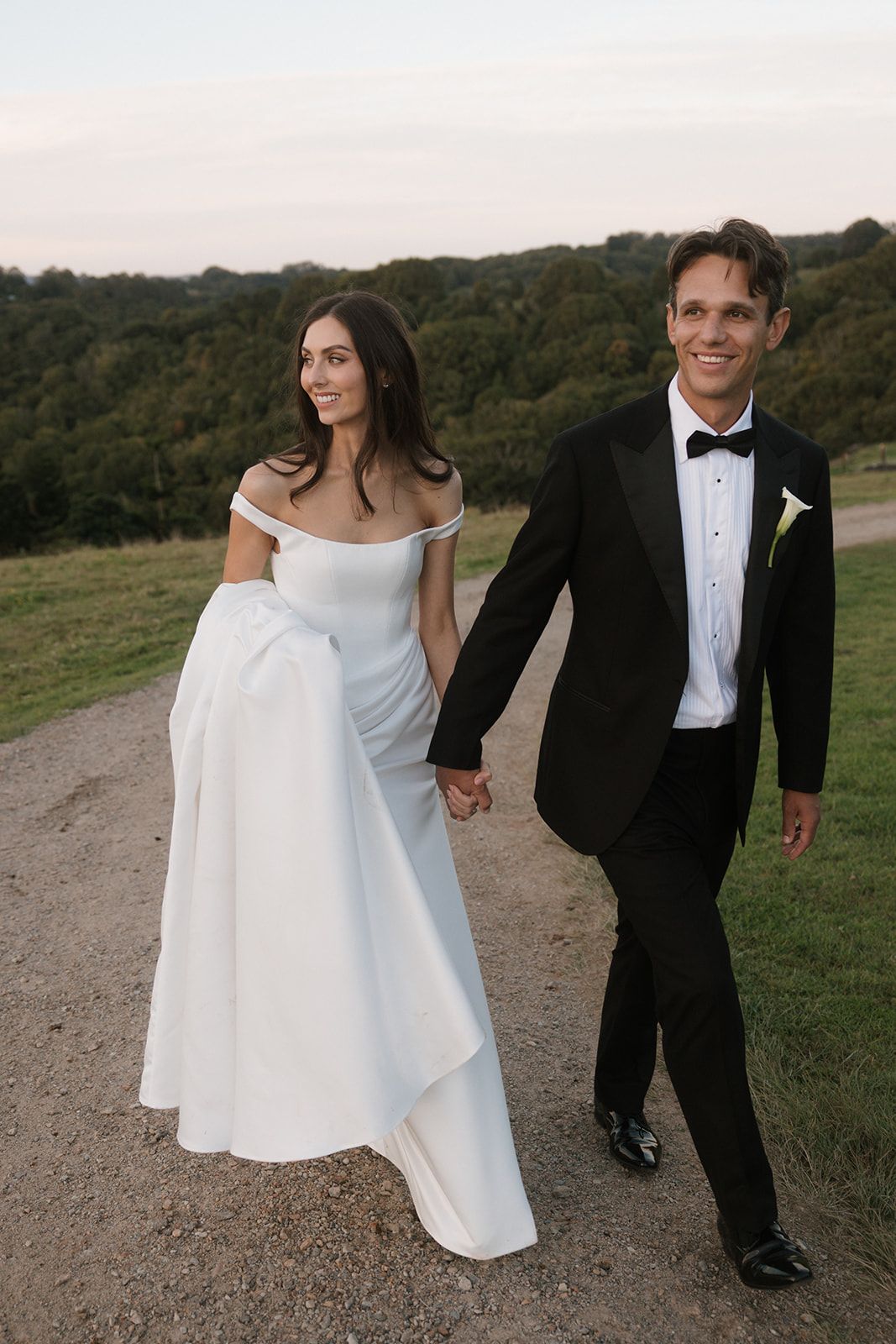 Bride in an off-the-shoulder white wedding gown holds hands with a groom in a black tuxedo while walking along a countryside path at sunset.