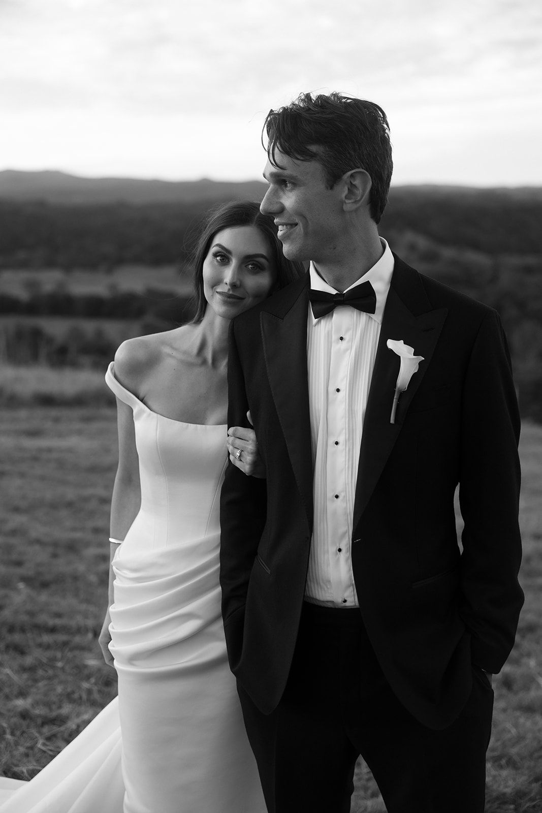 Black and white wedding portrait of a bride in an off-shoulder gown leaning on a smiling groom in a tuxedo and bow tie on a hillside.