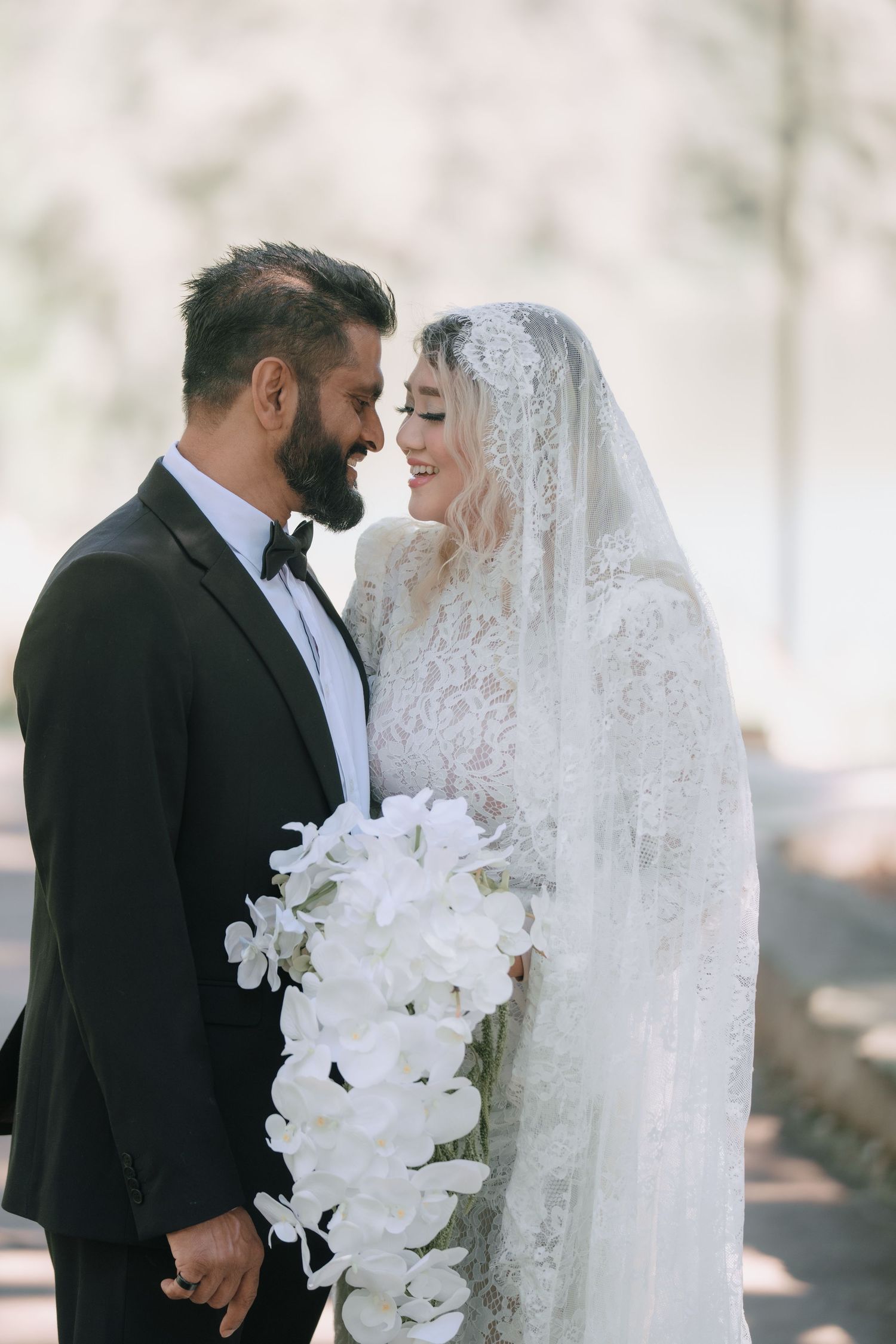 Smiling bride in a lace gown and long veil holding a white orchid bouquet while gazing at her groom in a black tuxedo and bow tie.