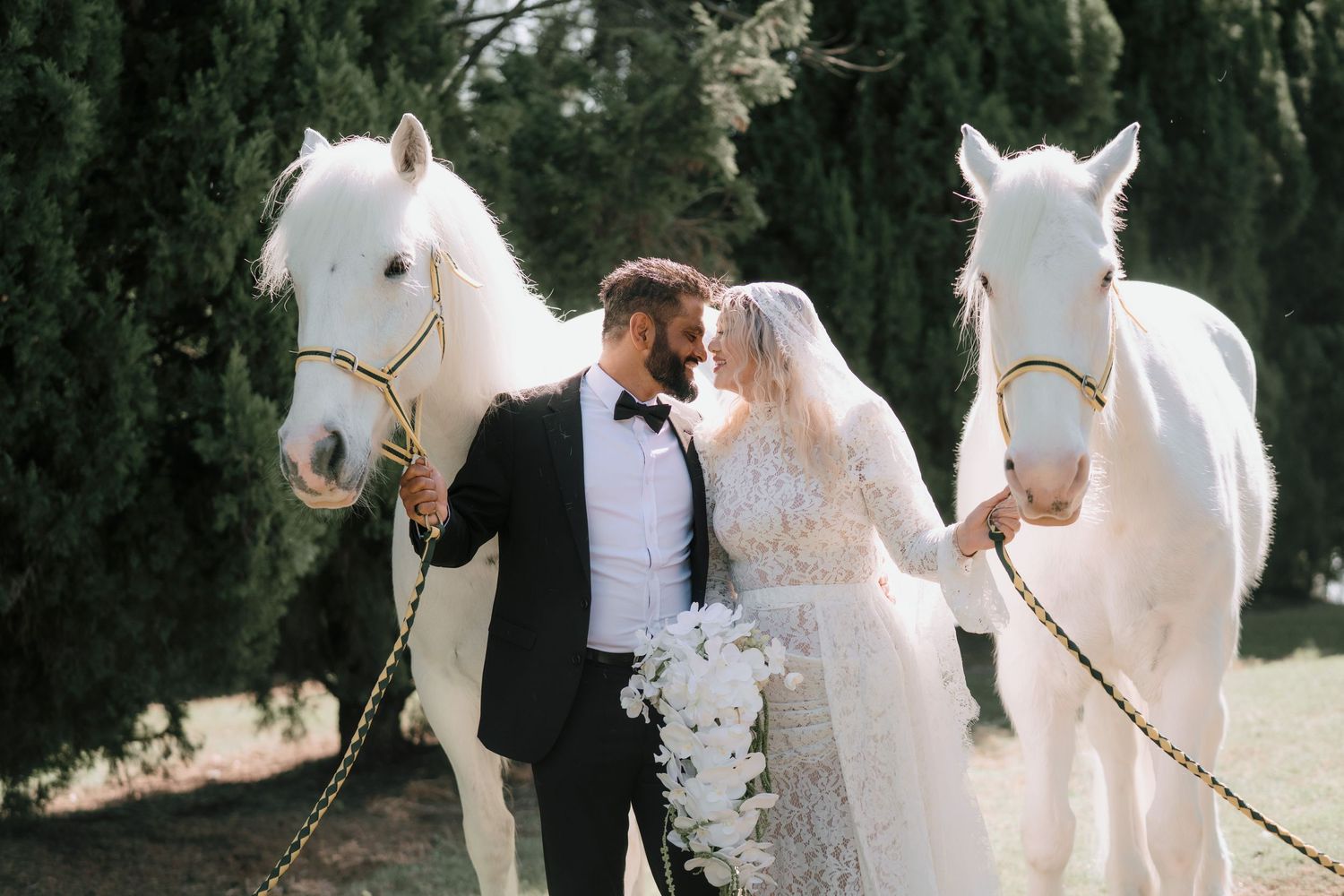 Bride and groom share a kiss between two white horses in a green forest, she holding a white bouquet, he in a black tuxedo with bow tie.