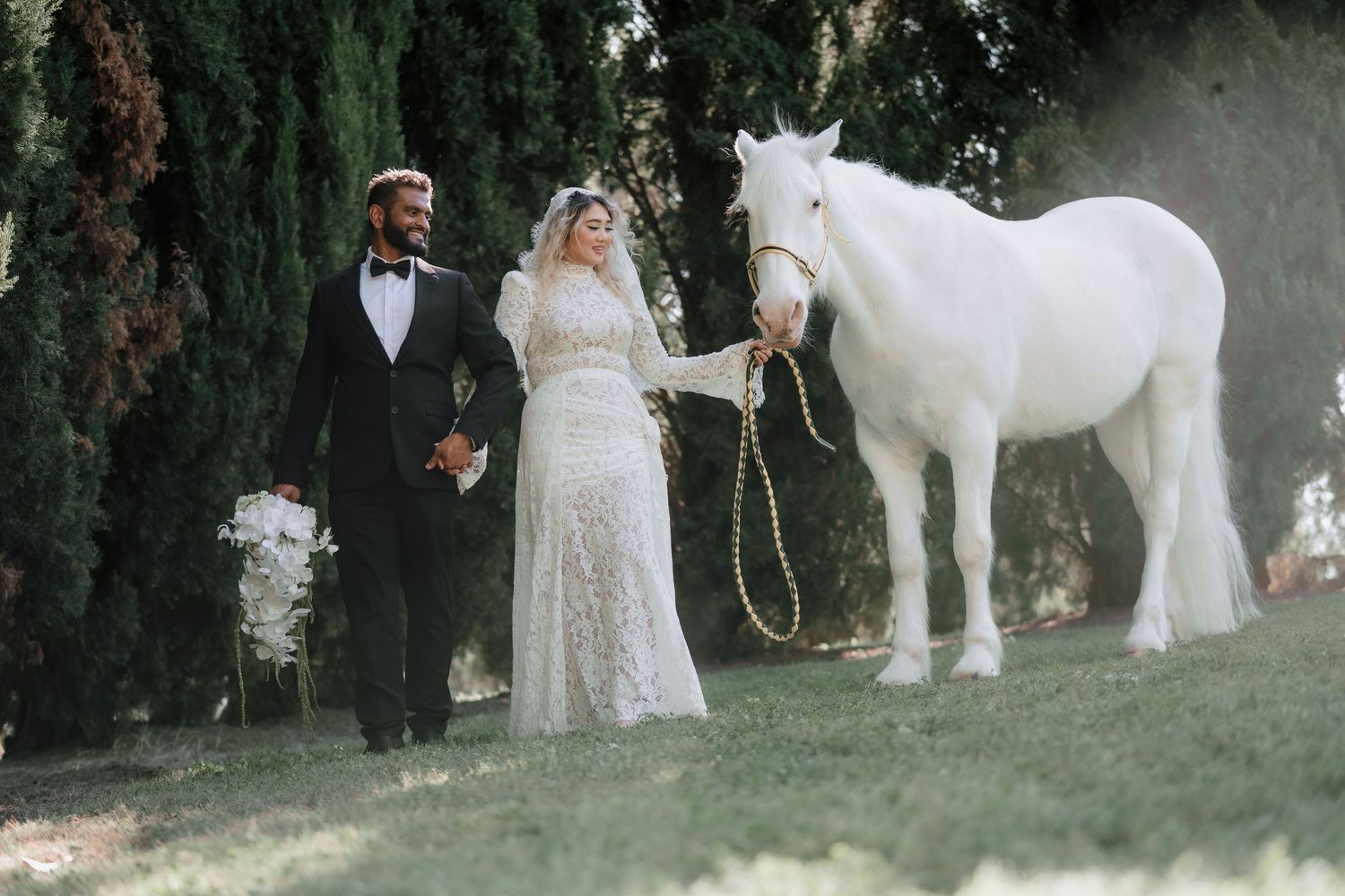 Bride in a lace gown and groom in a dark suit walk hand in hand with a white horse through a sunlit garden lined with tall green trees.