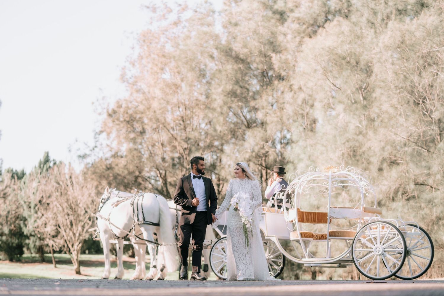 Bride in a white gown and groom in a tuxedo stand beside a white horse-drawn wedding carriage in a sunlit park surrounded by trees.