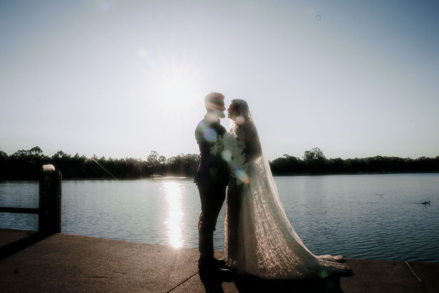 Bride in a lace wedding gown and groom in a suit embrace on a lakeside pier, backlit by a glowing sunset over the calm water.