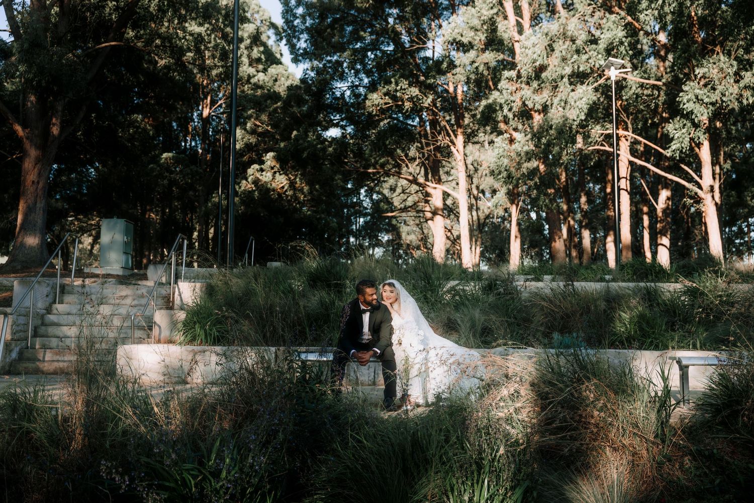 Bride in a white gown and groom in a dark suit sit together on a stone ledge, surrounded by tall eucalyptus trees and native garden plants.