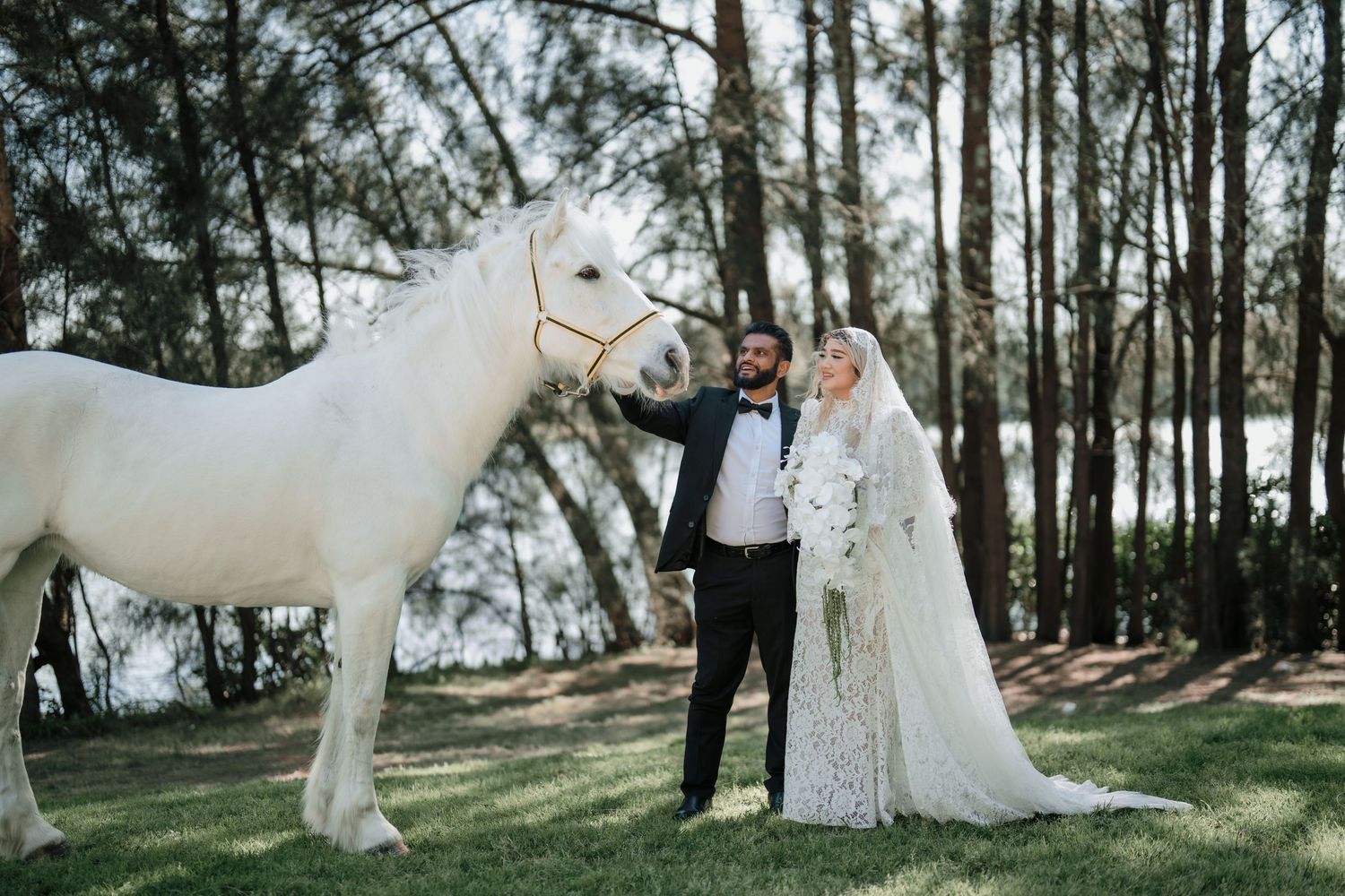 Bride in a flowing lace gown and veil stands with her groom beside a white horse on a grassy clearing surrounded by tall trees.