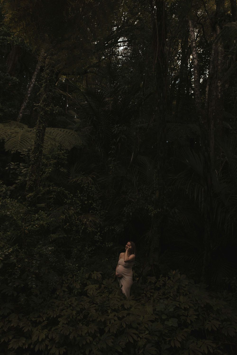 Pregnant woman in a light dress stands in a dark, dense tropical forest, surrounded by lush ferns and tall trees under dim moonlit light.