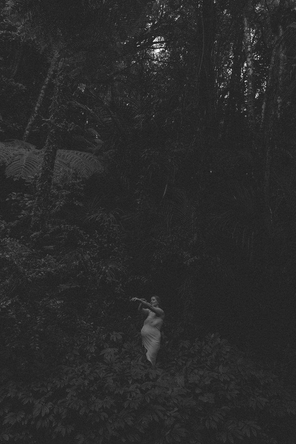 Black and white photo of a figure in a light dress standing in a dense, dark forest surrounded by ferns and thick tropical foliage.