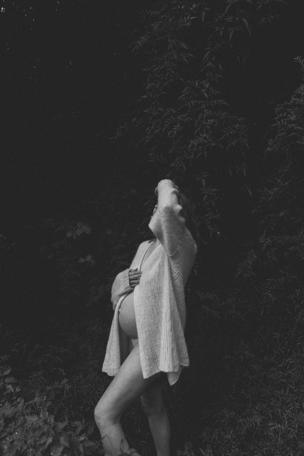 Black and white maternity portrait of a pregnant woman in a loose knit cardigan, head tilted back, standing among dark forest foliage.