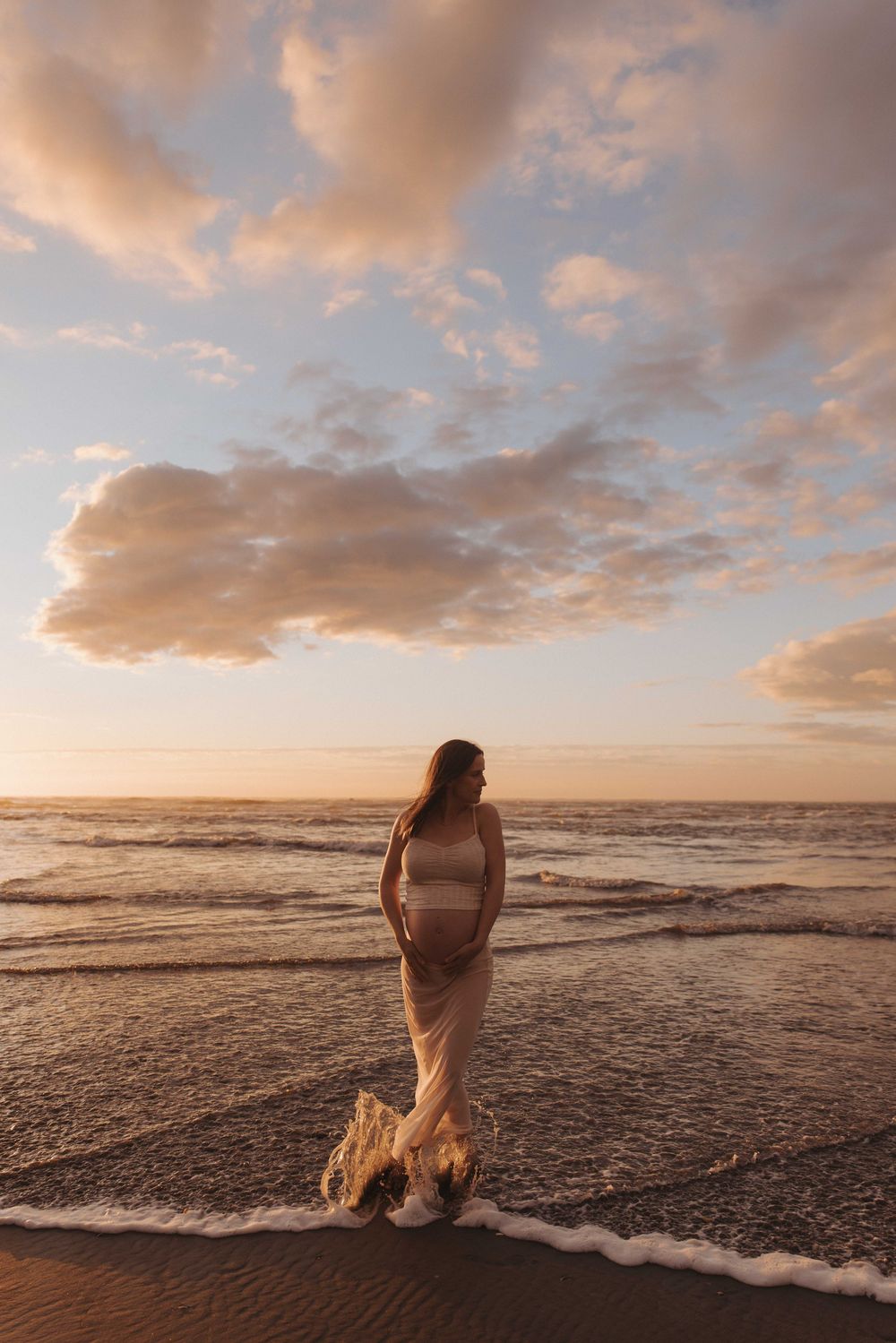 Pregnant woman in a flowing cream skirt stands at the ocean's edge during a golden sunset, with soft waves and glowing clouds behind her.
