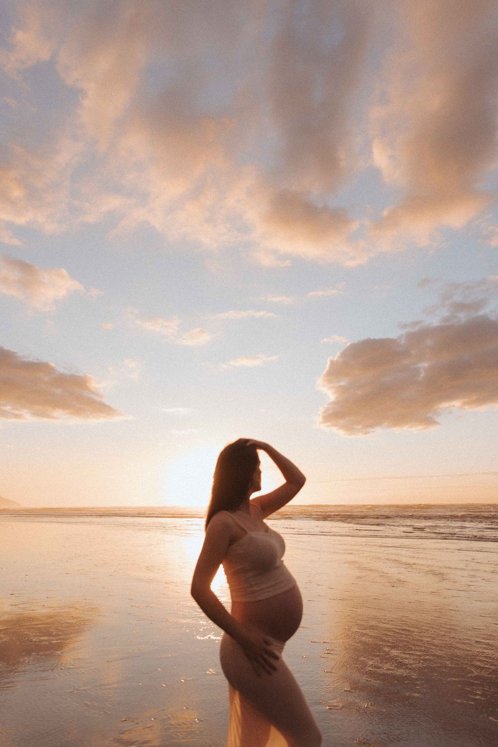 Pregnant woman in a light two-piece outfit stands on a wet sandy beach at sunset, one hand in her hair, under a soft pastel cloudy sky.