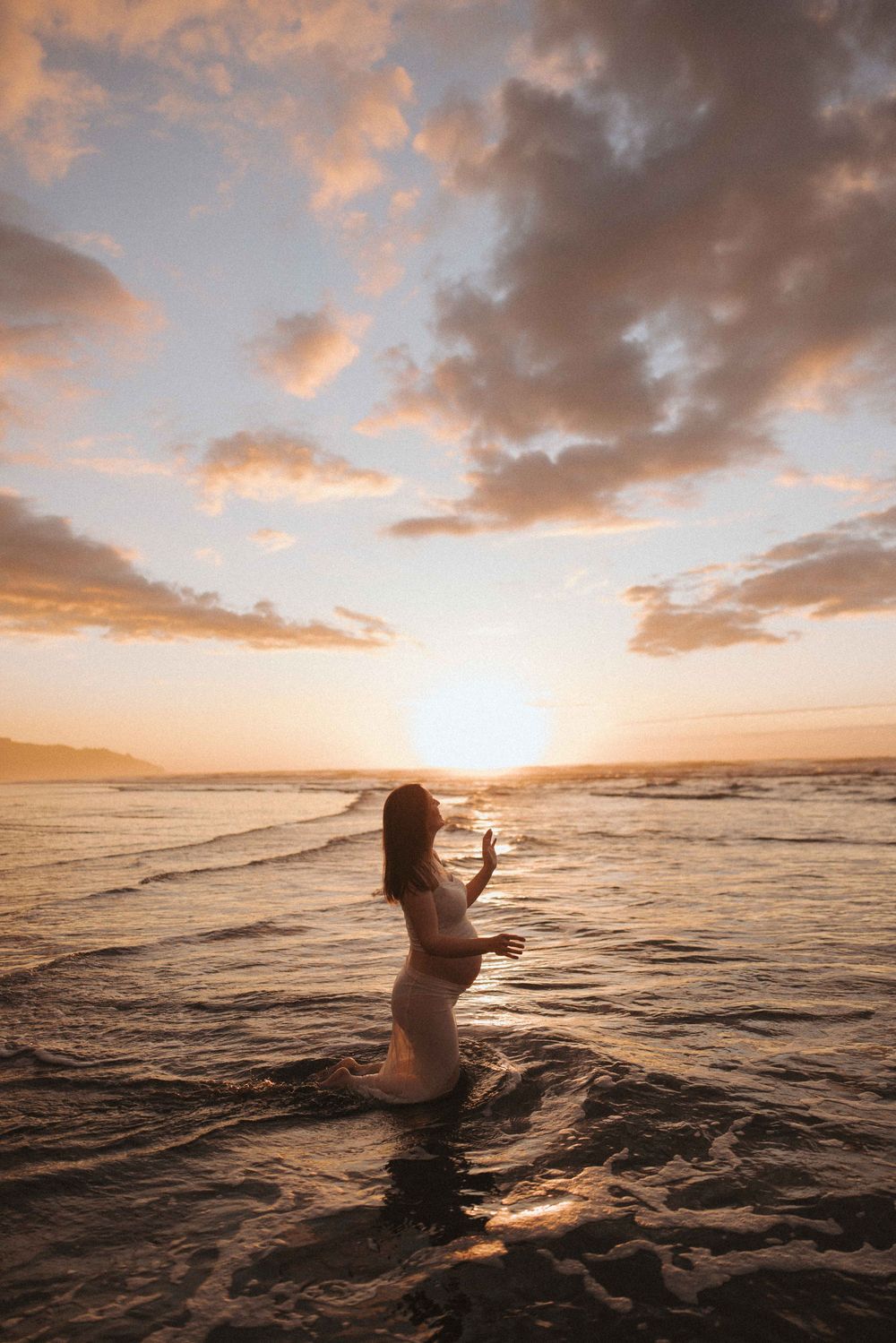 Woman in a white dress stands in shallow ocean waves at golden sunset, with dramatic clouds and warm light reflecting on the water.