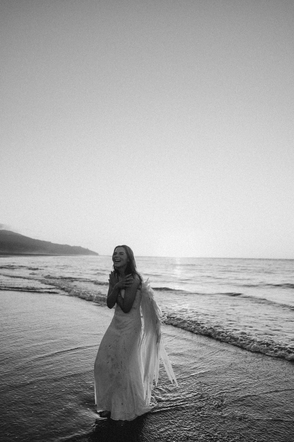 Black and white photo of a smiling woman in a flowing white dress with angel wings standing in shallow ocean waves at sunset.