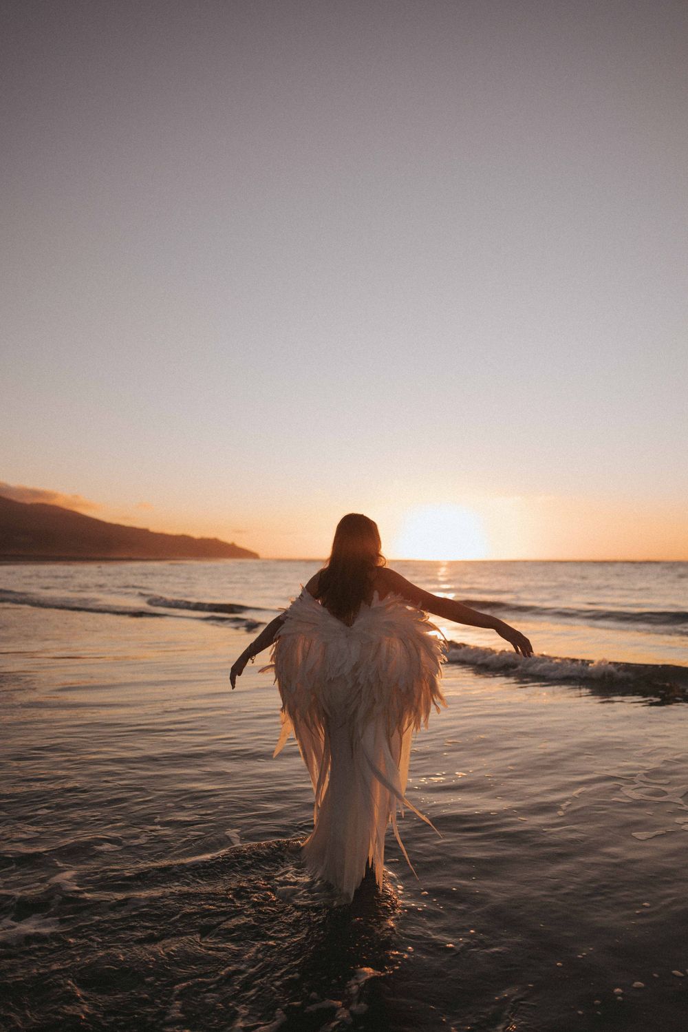 Woman in a white feathered angel wing costume wades into calm ocean waves with arms outstretched at a golden sunset.