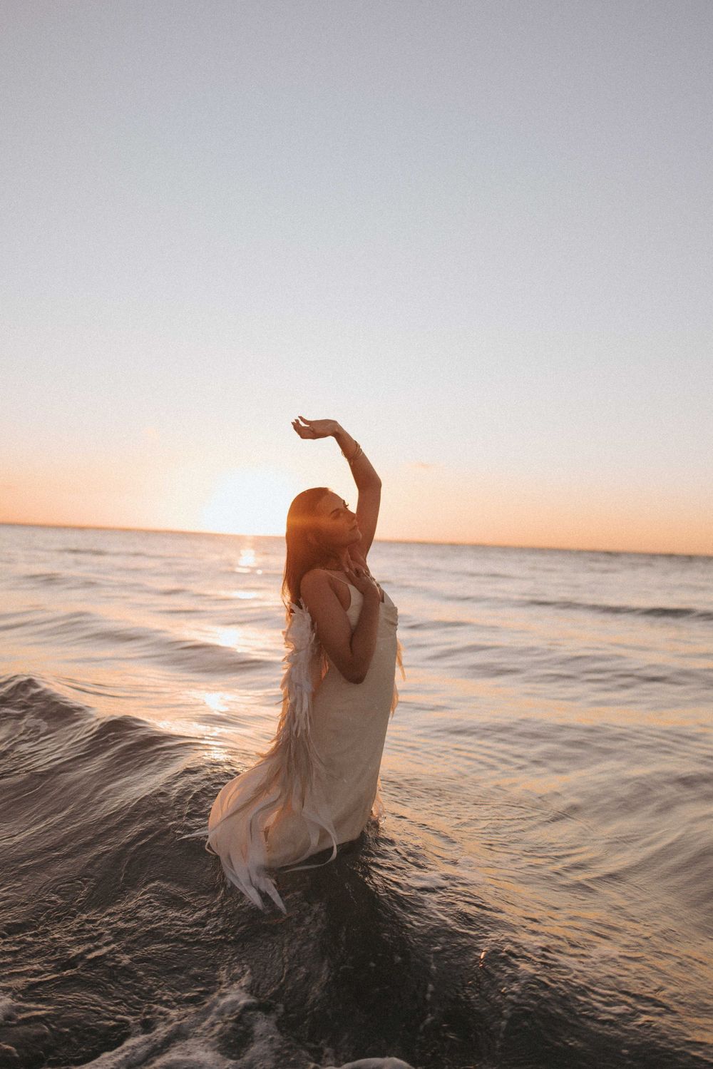 Woman in a flowing white dress stands in ocean waves at sunset, one arm raised toward the sky as golden light reflects on the water.