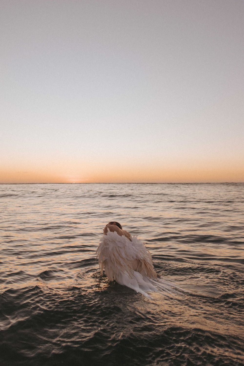 Person in a flowing white feathered gown wading into the calm ocean at sunset, with a soft peach and pink sky over the horizon.