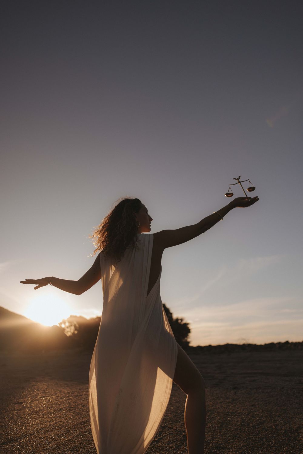 Silhouette of a woman in a flowing white dress holding small balance scales in her outstretched hand against a glowing sunset sky.