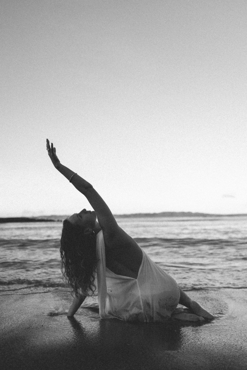 Black and white photo of a woman in a flowing dress sitting on the beach at the water's edge, one arm stretched toward the sky.