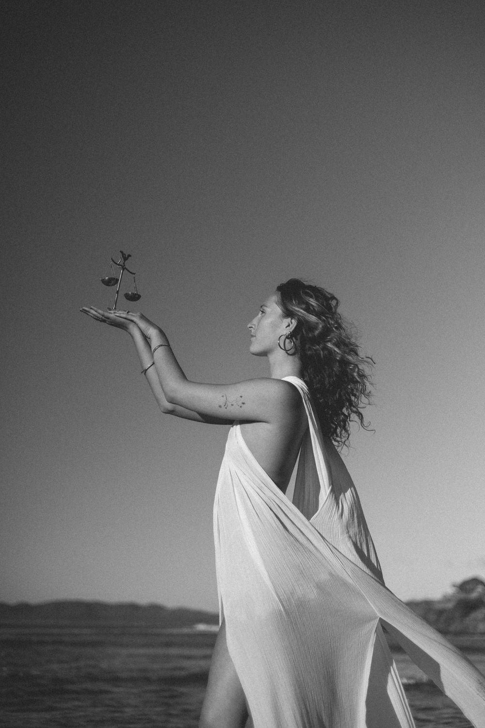 Black and white photo of a woman in a flowing white dress holding a small plant sprig in her raised hand on a beach by the sea.
