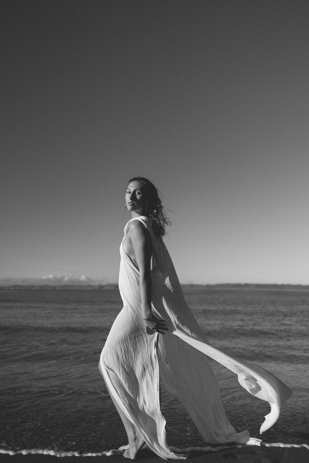 Woman in a flowing white dress walks along the ocean shoreline, glancing over her shoulder in a black and white beach portrait.