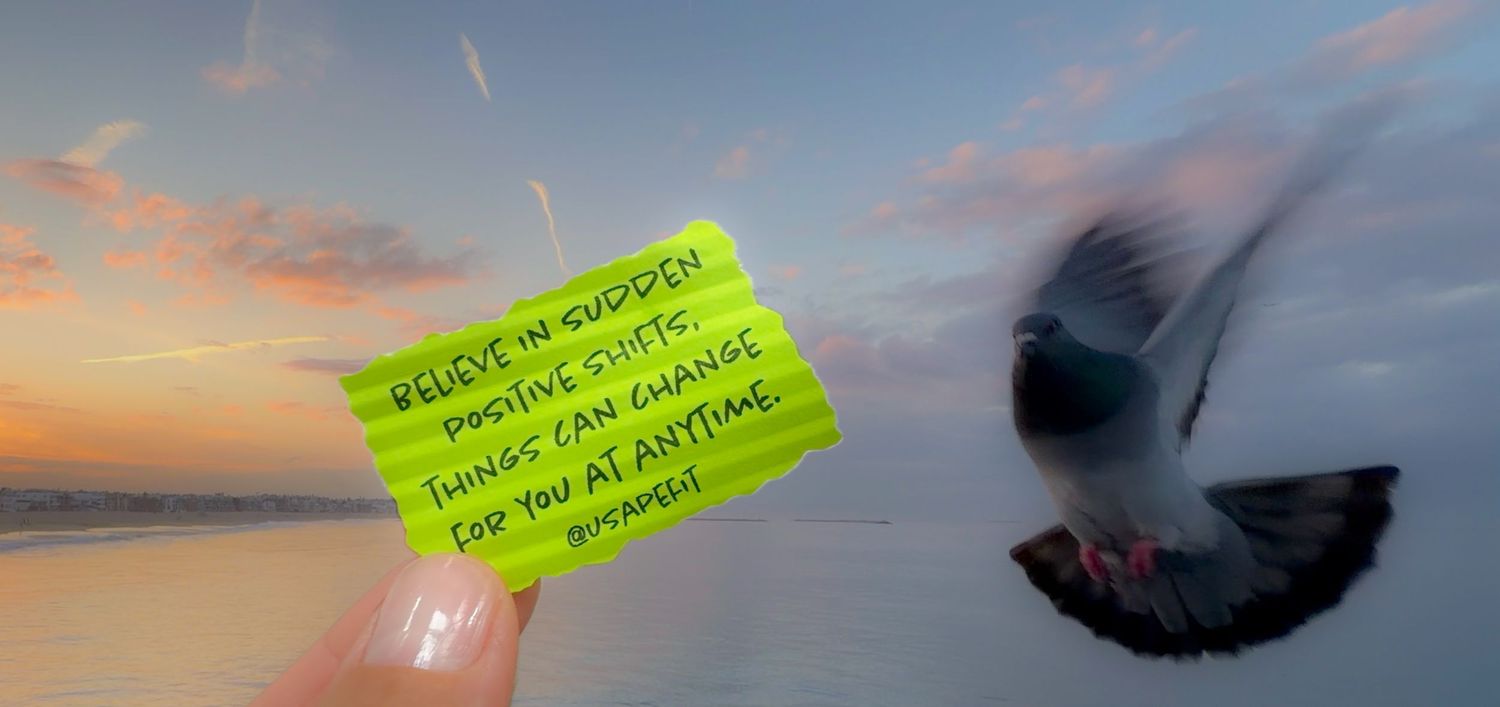 A hand holds a green sticky note with an inspirational message as a pigeon flies past a pink and blue sunset sky.