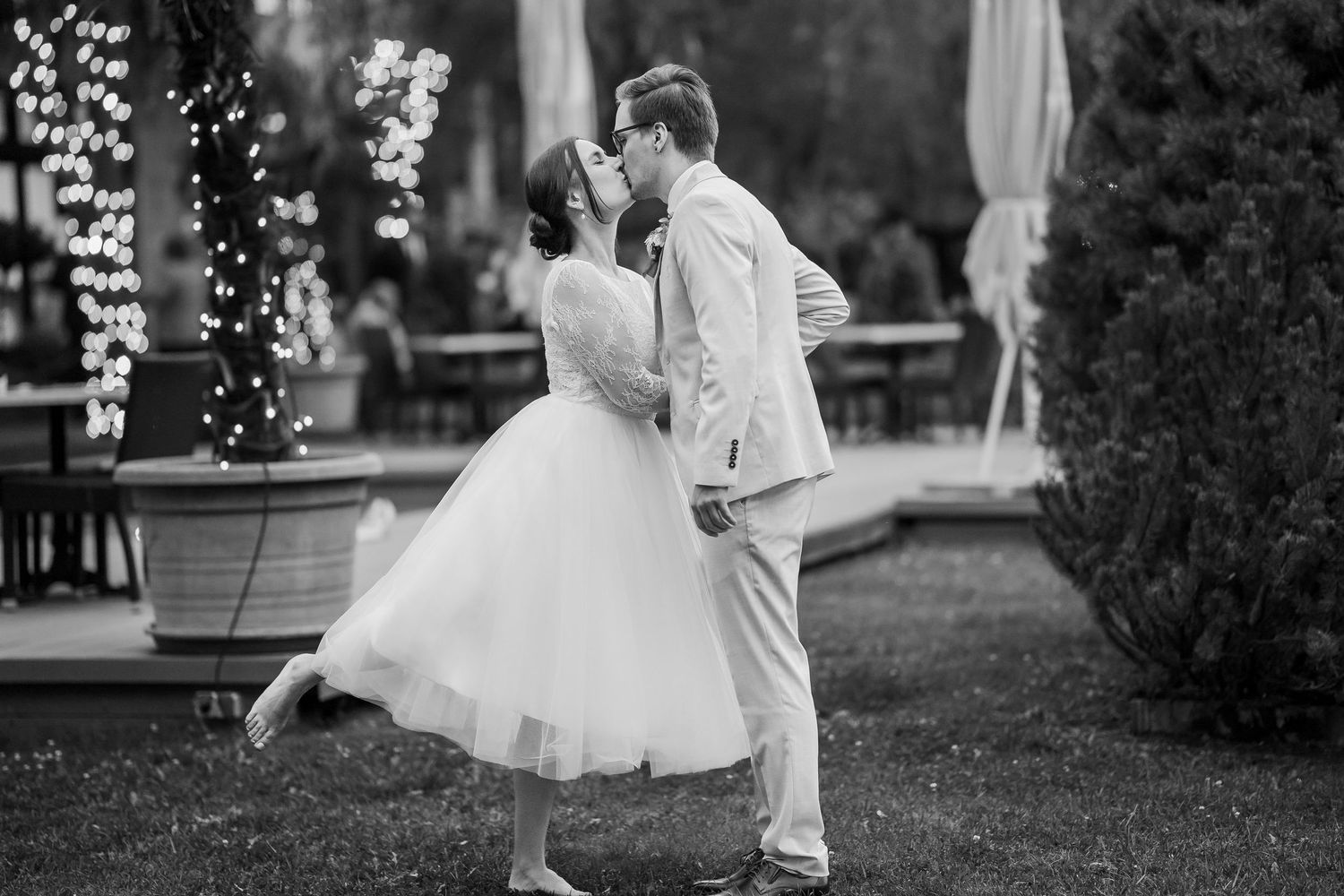 Black and white photo of a bride in a tulle dress and a groom in a suit kissing outdoors beside string lights and potted greenery.
