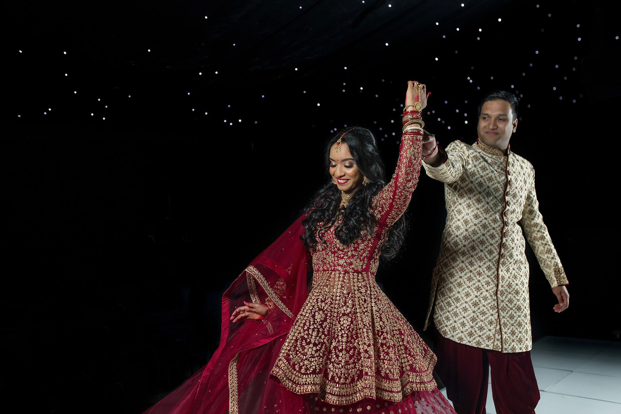 Couple in traditional Indian wedding attire dancing elegantly against a dark backdrop with sparkling lights.