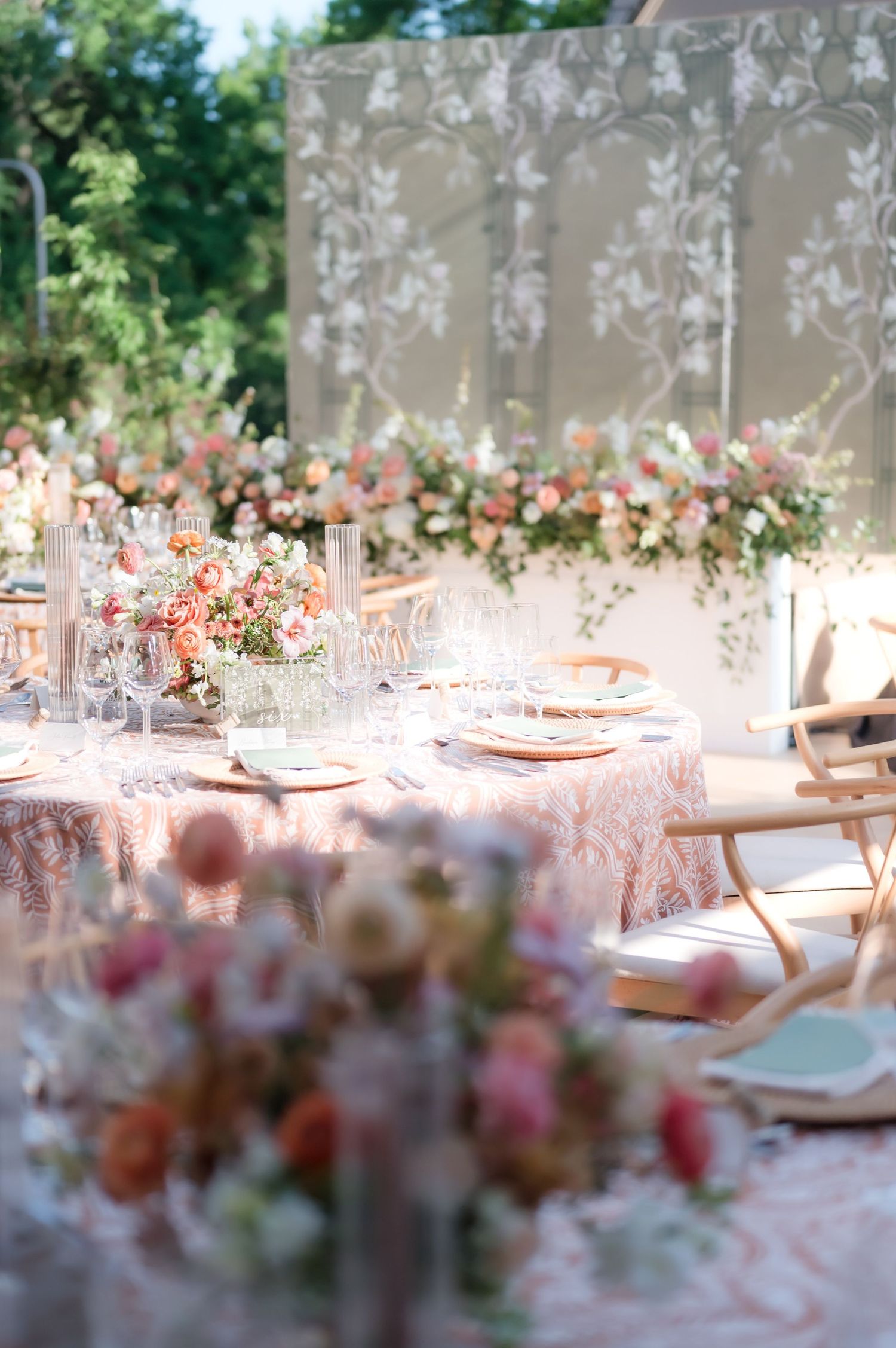 Elegant outdoor wedding reception table with pink floral centerpieces, lace tablecloth, and floral backdrop.