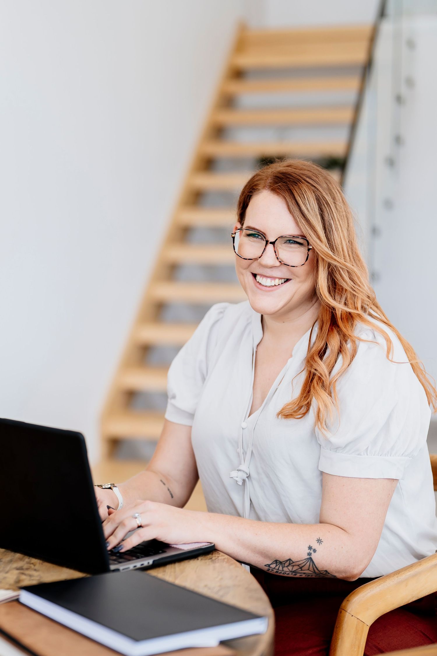 Smiling redhead woman with glasses working on laptop at desk near wooden staircase in bright home office headshot photographer queensland