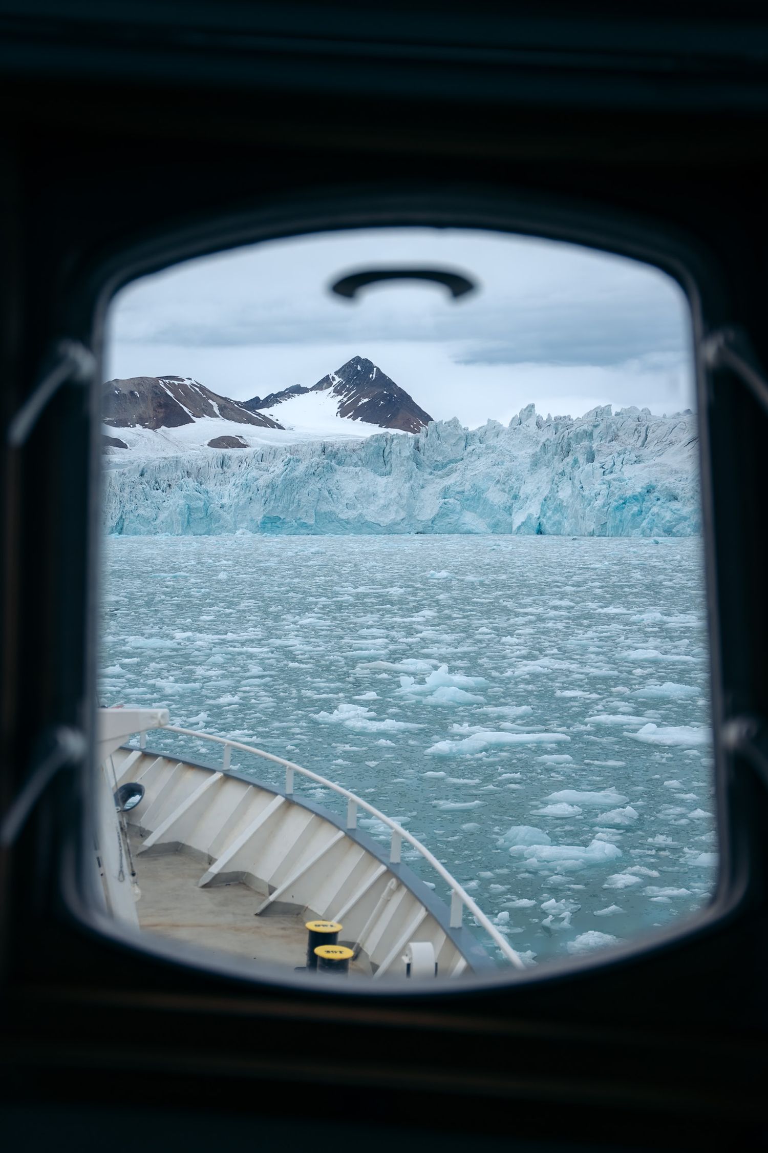 Front glaciaire cadré dans un hublot sombre du Polarfront, proue visible en bas